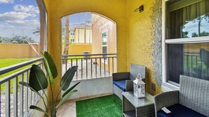 Un balcon donnant sur un immeuble jaune, un canapé en osier, une petite table avec une lanterne et une plante en pot.