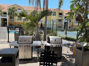 En grillplats med Weber-grillar, ett poolområde med uppsatta regler och en tennisbana i bakgrunden.
