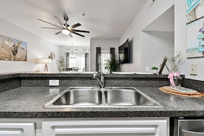 A kitchen with a double sink, granite countertop, ceiling fan, and a dining area in the background.