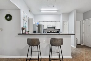 A modern kitchen with white cabinets, a stainless steel refrigerator, and a black countertop. There are two bar stools and a small potted plant on the counter. - Ground floor 2 Bedroom One Bathroom Condo in gated community with Pool (Seminole)