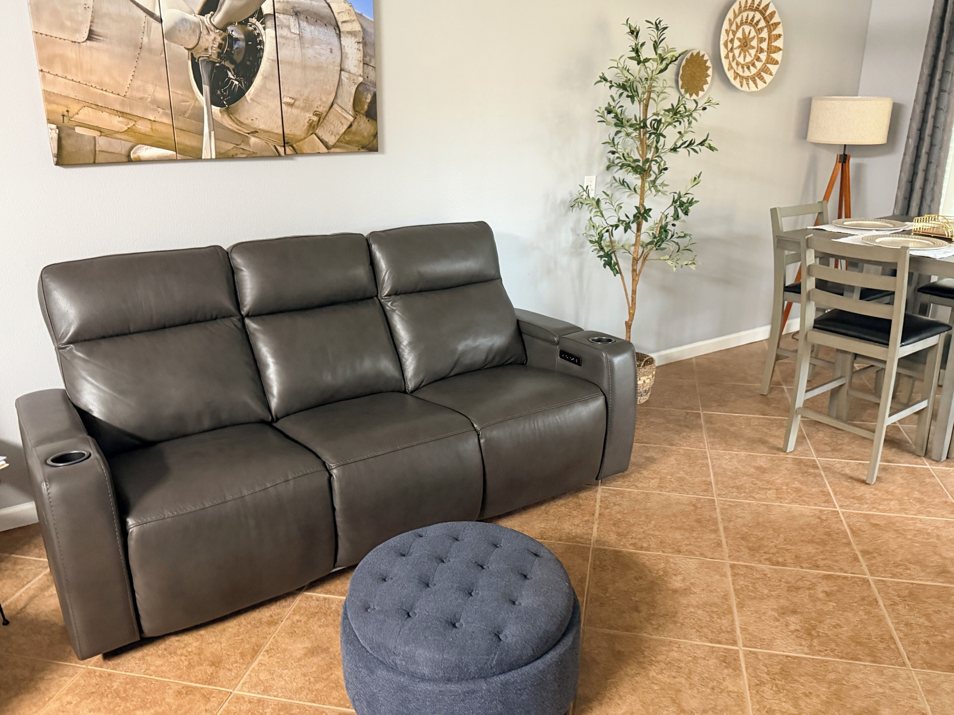 A dark brown leather sofa with a tufted ottoman in front, a dining table with chairs, a potted plant, and a wall-mounted artwork.