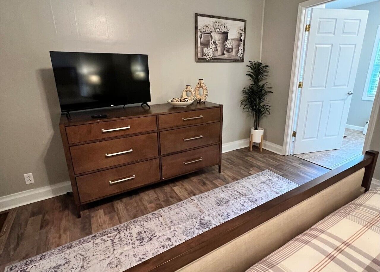 A wooden dresser with drawers, a flat-screen TV on top, a patterned rug, and a door in the background.