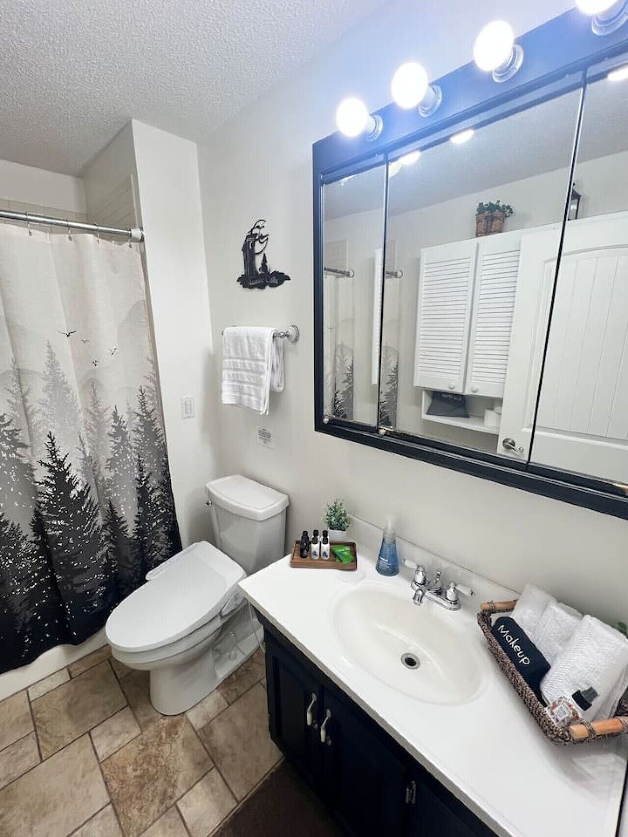 Een badkamer met douchegordijn met een boslandschap, toilet, wastafel met toiletartikelen en spiegel met verlichting.
