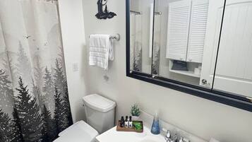Een badkamer met douchegordijn met een boslandschap, toilet, wastafel met toiletartikelen en spiegel met verlichting.