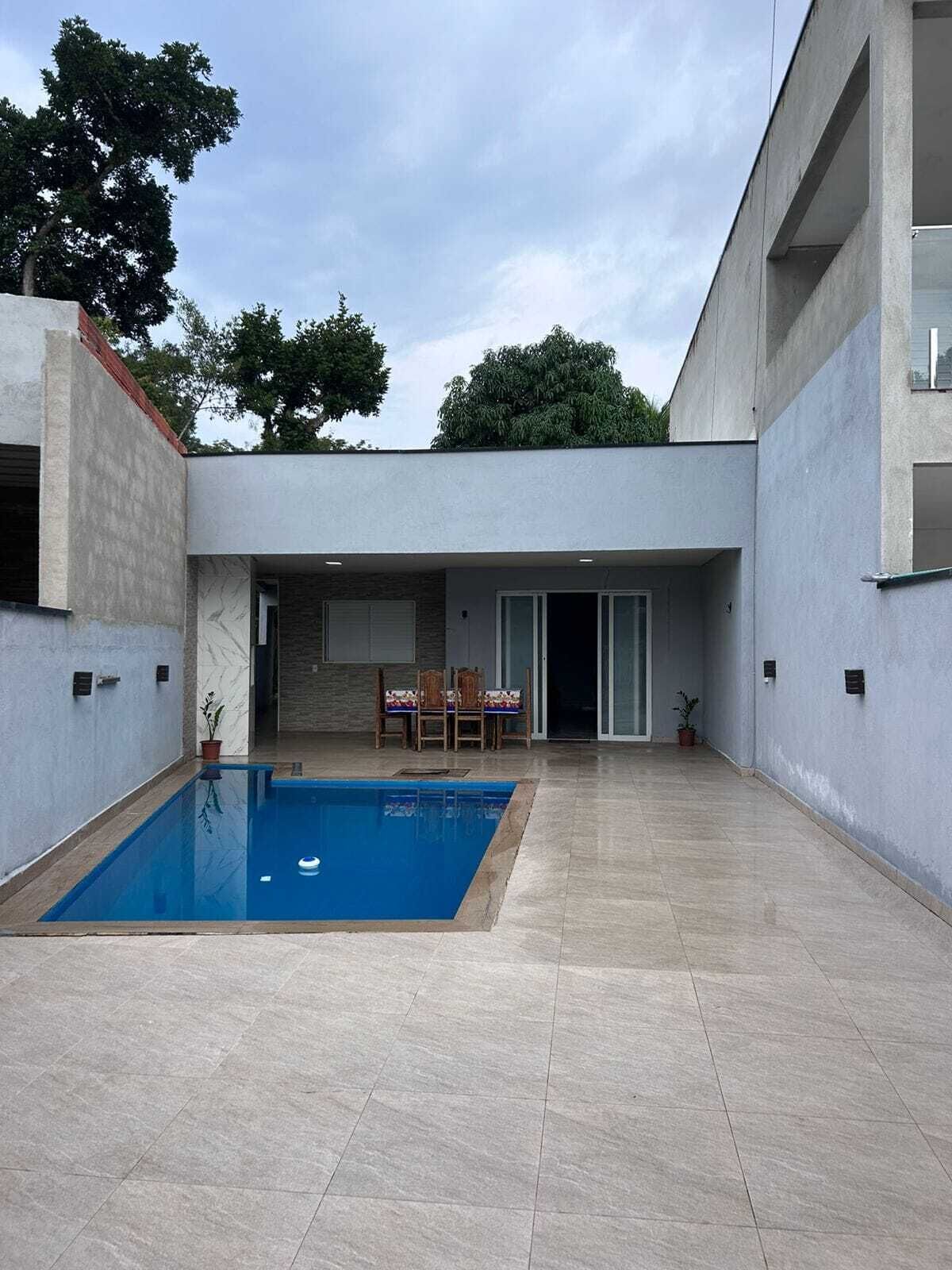 Uma área moderna com piscina ao ar livre, de águas cristalinas e azuis, um espaço de estar coberto com cadeiras, e um edifício com telhado plano.