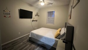 A bedroom with a bed, a television mounted on the wall, a ceiling fan, and a window with blinds.