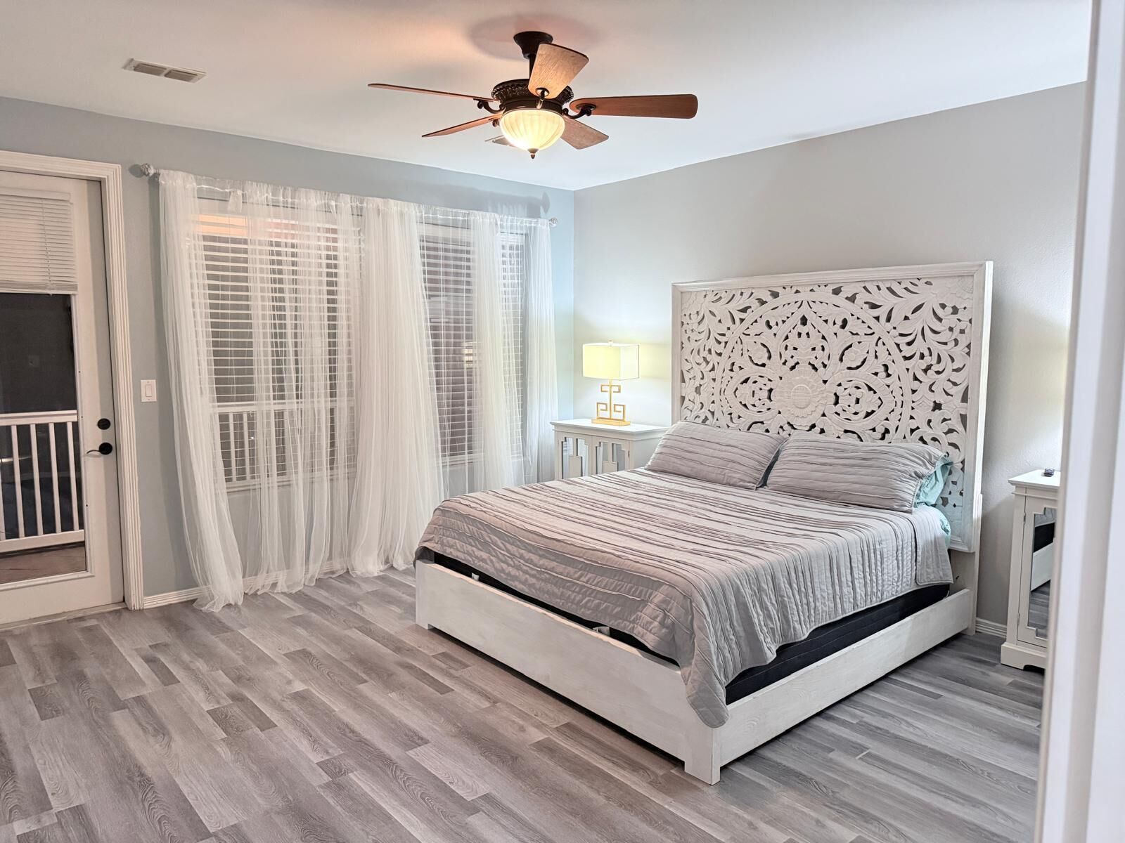 A bedroom with a bed, a ceiling fan, a window with sheer curtains, and a door.