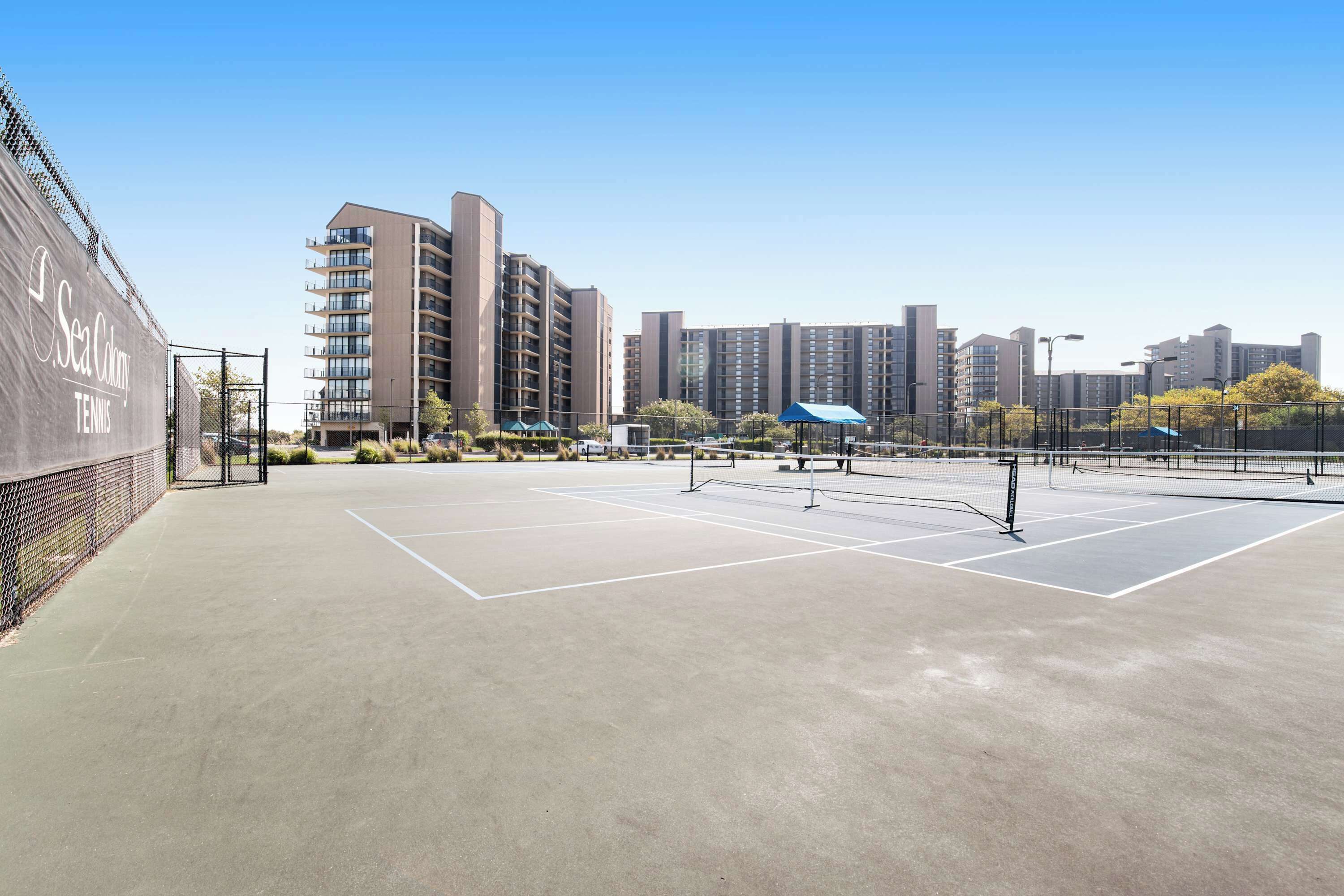 A tennis court with a net, surrounded by a fence, and adjacent to a building labeled 'Sea Colony Tennis.'