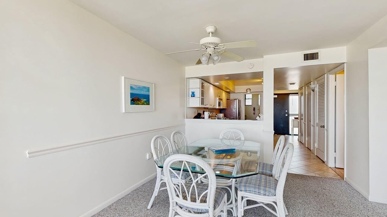 A dining area with a glass table and white chairs, a ceiling fan, and a kitchen visible through an open door.