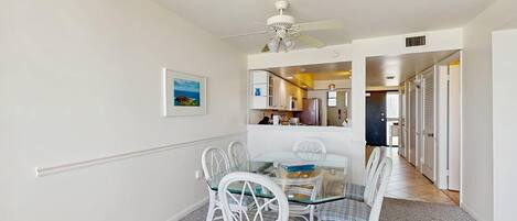 A dining area with a glass table and white chairs, a ceiling fan, and a kitchen visible through an open door.