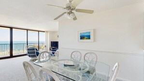 Una habitación con una mesa de vidrio, sillas blancas, un ventilador de techo, un ventanal con vista al mar y un cuadro en la pared.