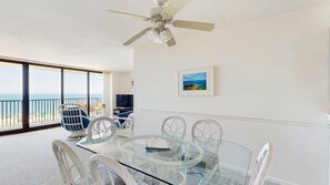 A room with a glass table, white chairs, a ceiling fan, a large window with a view of the sea, and a framed picture on the wall. - Sea Colony - 603N Edgewater House (Bethany Beach)
