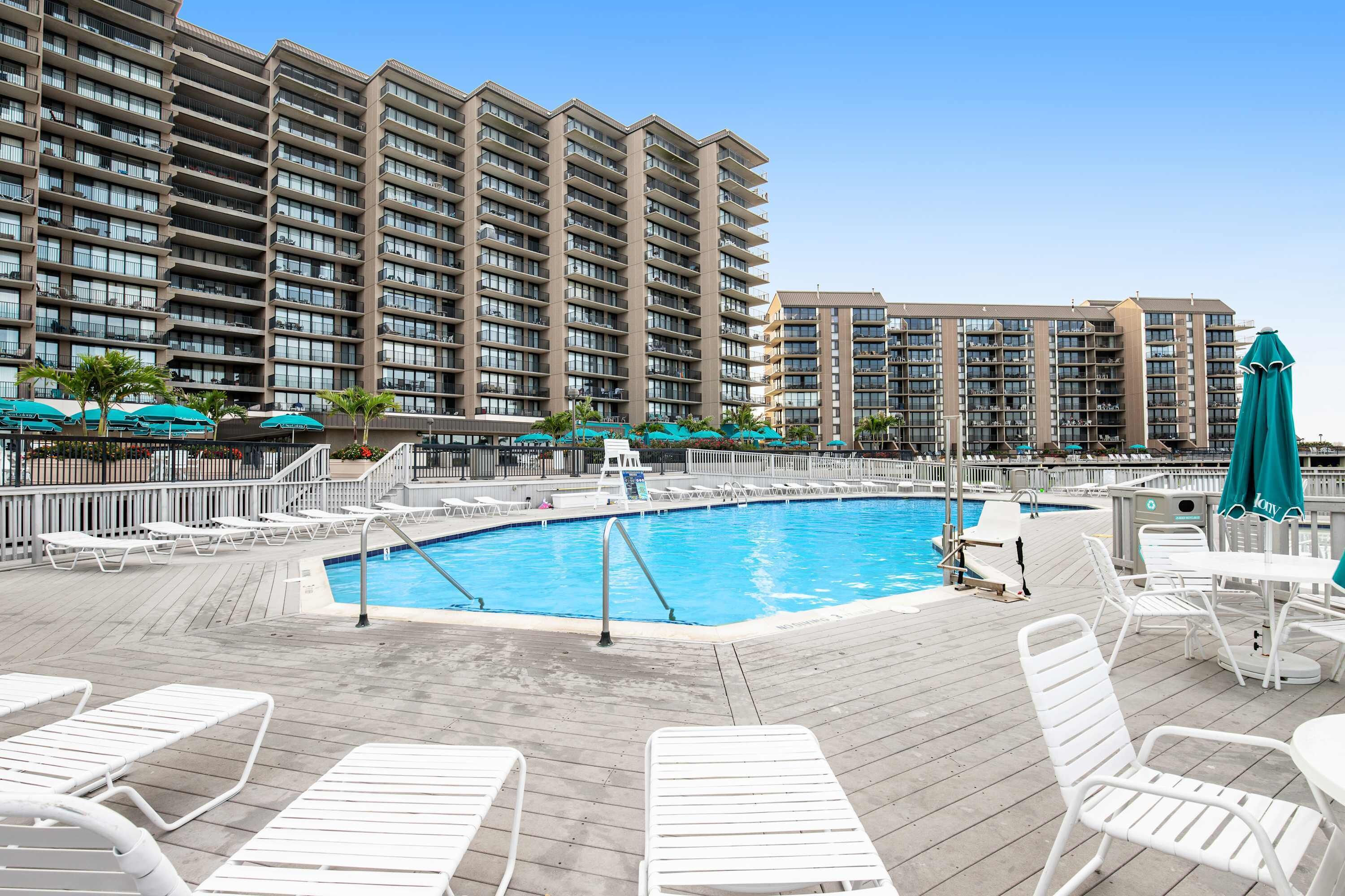 A hotel pool area with multiple lounge chairs, a swimming pool, and high-rise buildings in the background.