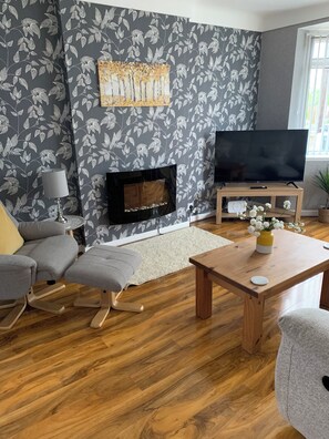 En stue med et fladskærms-tv, pejs, gråt blomstret tapet, et træ sofabord og en grå sofa.