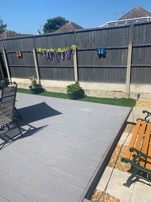 Eine Holzterrasse mit einem Metalltisch und -stühlen, umgeben von einer niedrigen Mauer mit herabhängenden violetten Blumen und zwei Schmetterlingsdekorationen.