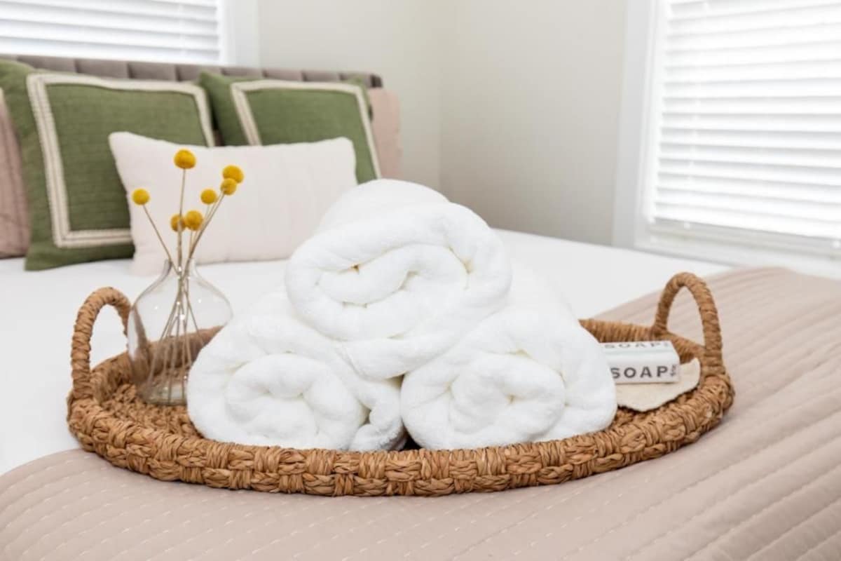 A neatly arranged set of white towels on a woven tray with a label, placed on a bed with a beige bedspread and pillows in the background.
