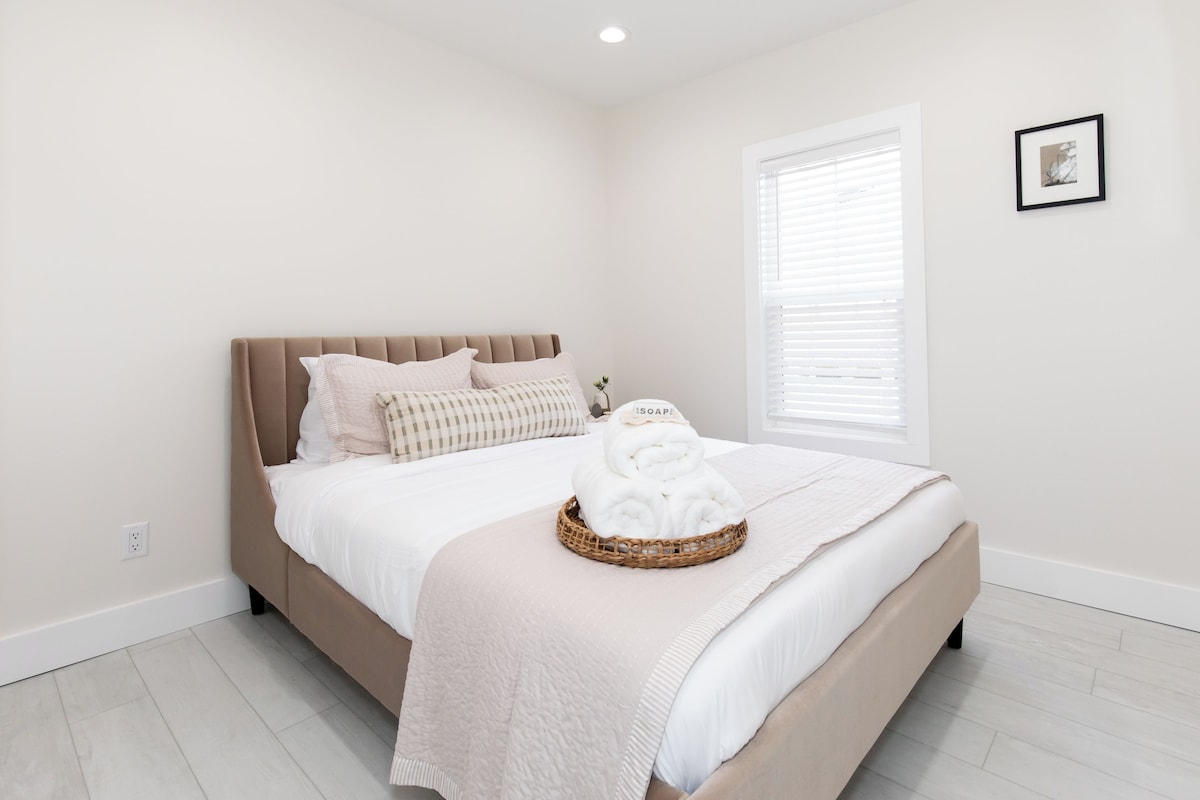 A bedroom with a bed, a bedside table, a window with blinds, and a wall-mounted picture.