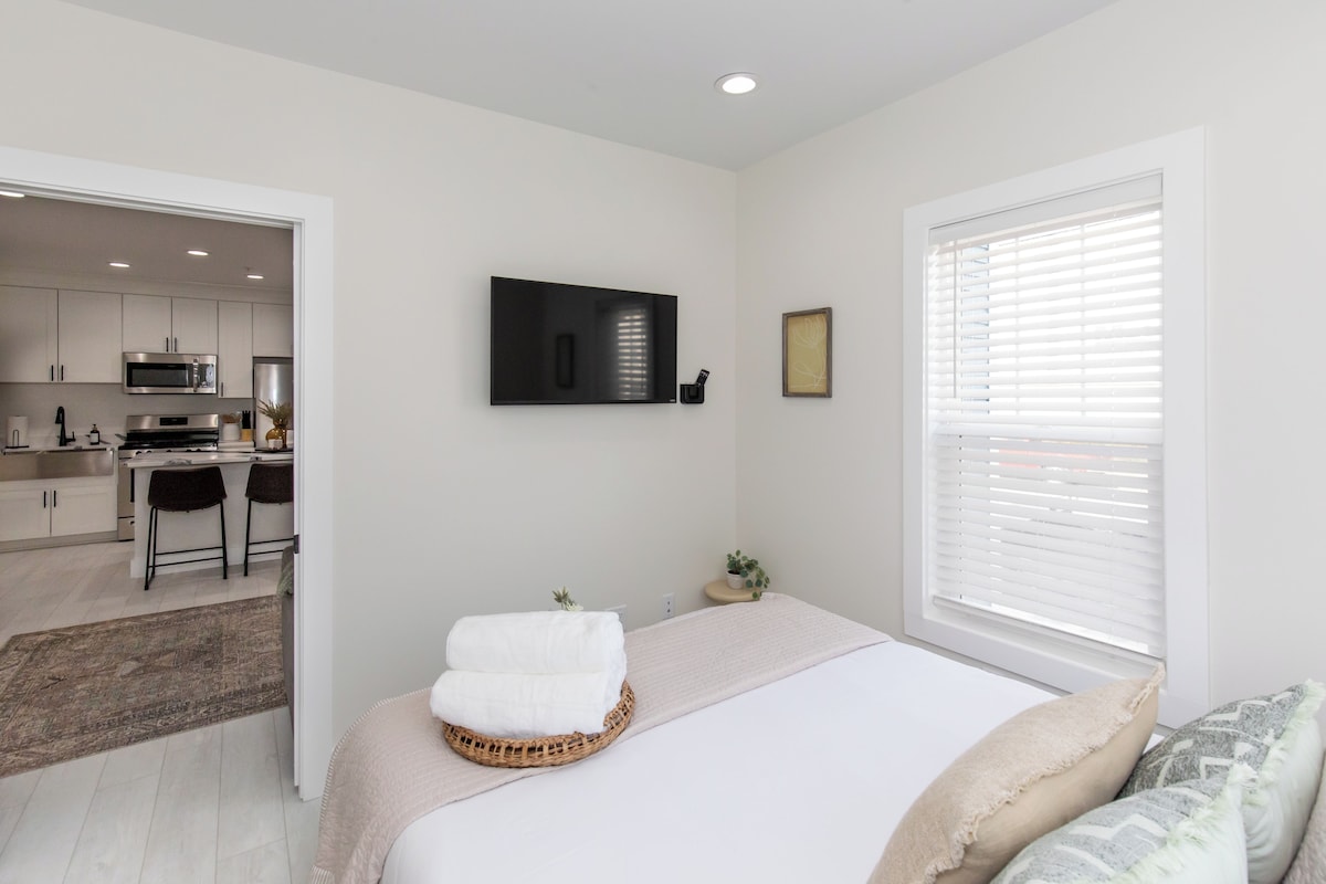 A modern bedroom with a bed, a TV mounted on the wall, and a view of the kitchen through an open door.