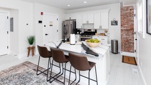 Una cocina moderna con armarios blancos, una isla central con taburetes, un refrigerador de acero inoxidable y una pared de ladrillo decorativa.