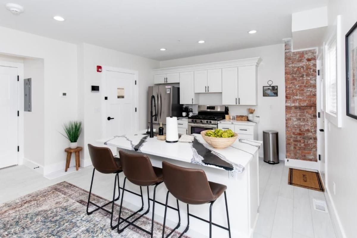 A modern kitchen with white cabinetry, a central island with bar stools, a stainless steel refrigerator, and a brick accent wall.