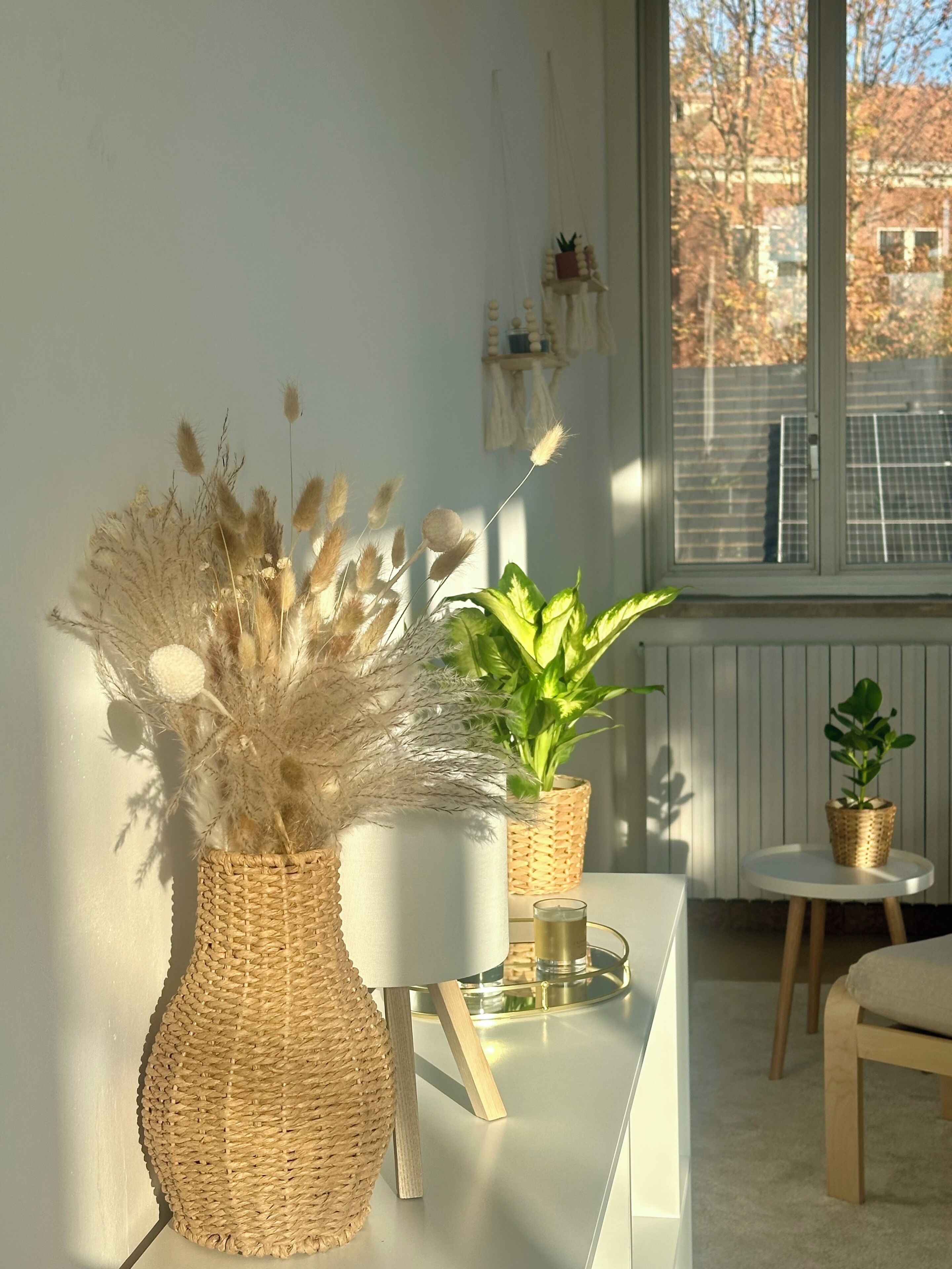 Una habitación con una mesa blanca, dos jarrones de mimbre con plantas, un vaso sobre la mesa y vistas de un edificio a través de una ventana.