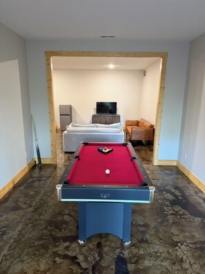 Uma mesa de bilhar com taco, um sofá e uma televisão em um ambiente com piso de concreto.