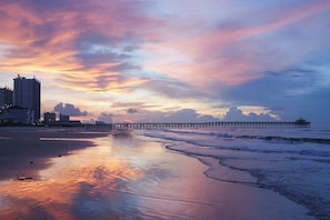 Ein Strand mit einem Steg, der ins Wasser führt, Gebäuden im Hintergrund und einem Sonnenuntergangshimmel.
