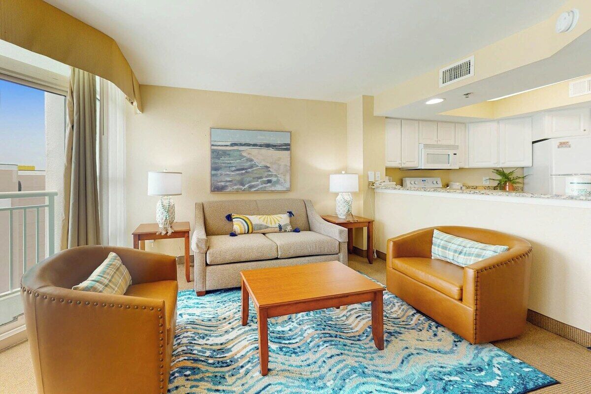 A hotel room with a sofa, armchairs, a coffee table, a kitchen area, and a painting on the wall.