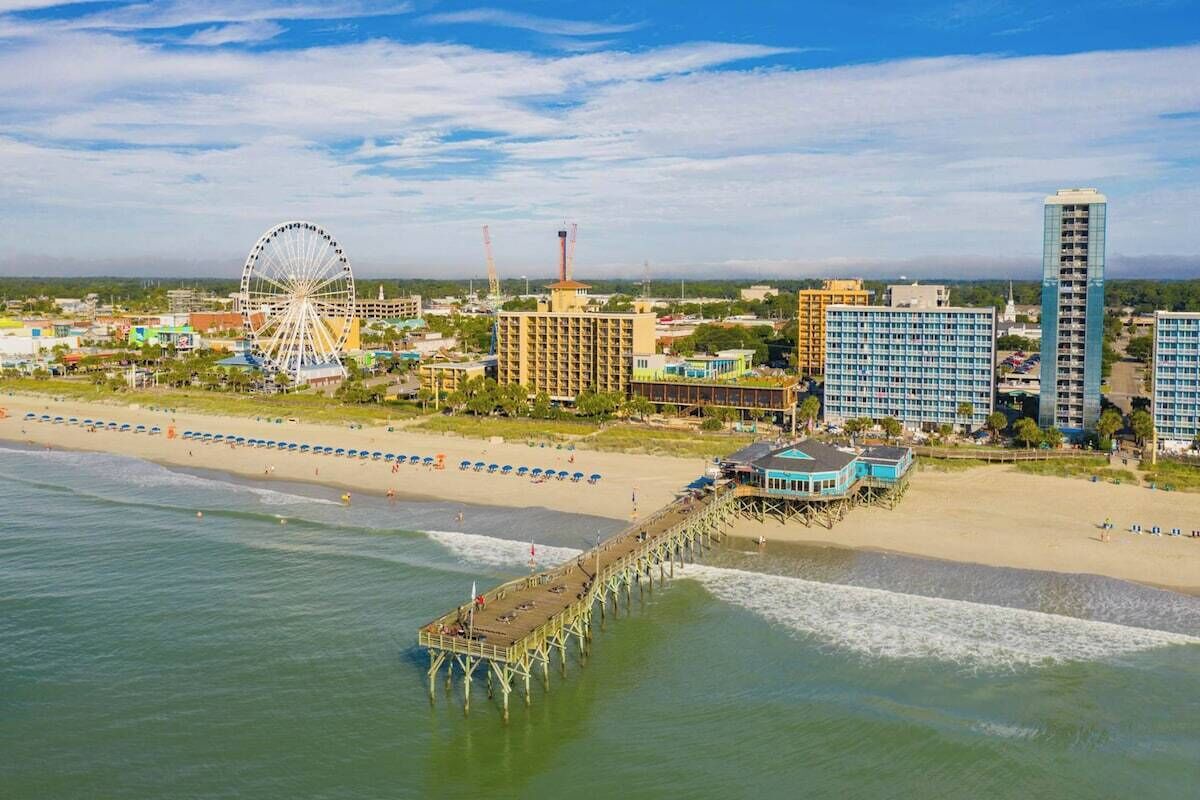 A coastal cityscape with a pier extending into the water, a large Ferris wheel, and high-rise buildings.