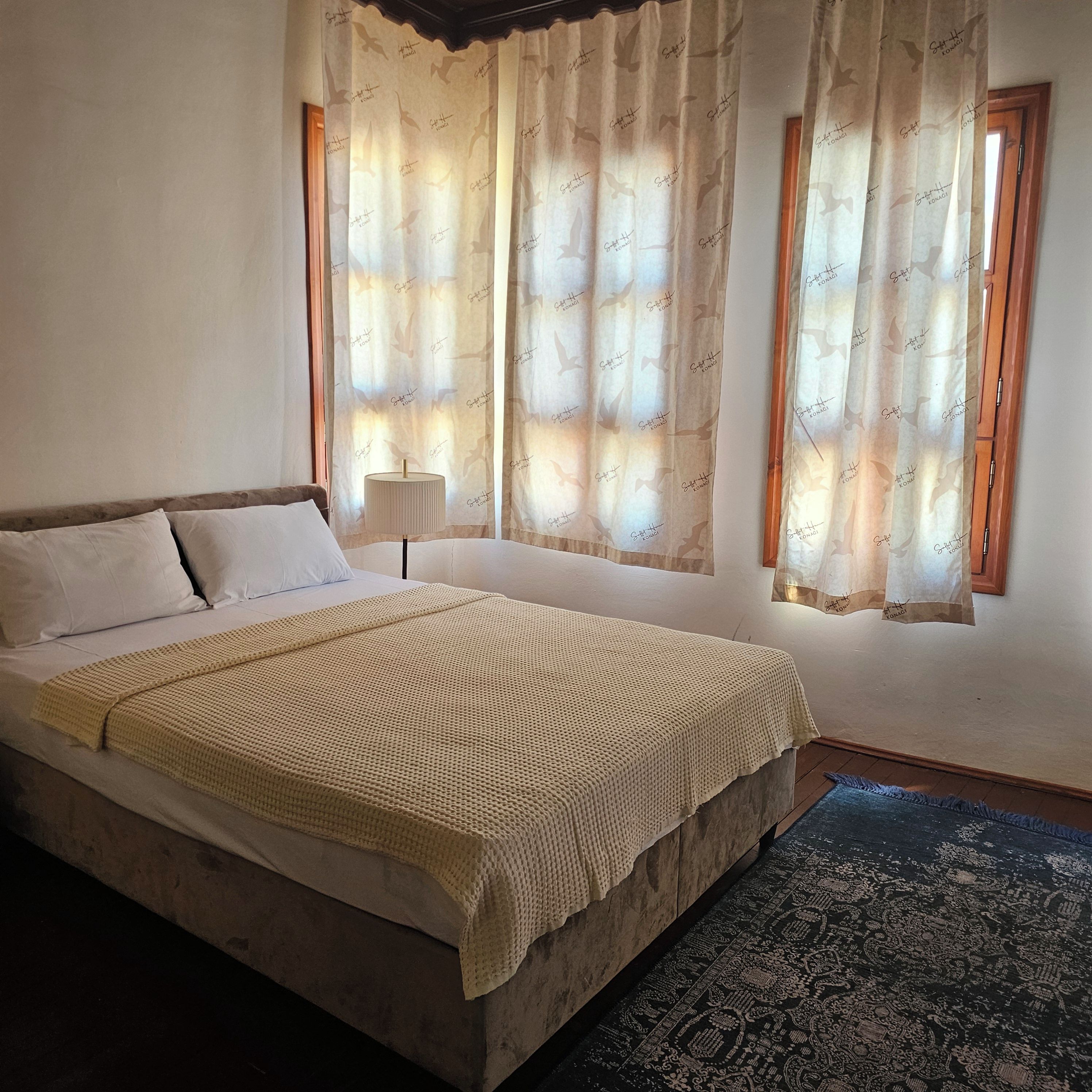 Un dormitorio con cama, cortinas, una lámpara y un tapete estampado.