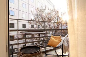 Un balcón con una silla de mimbre, una pequeña mesa redonda y vistas a un edificio con varias ventanas.