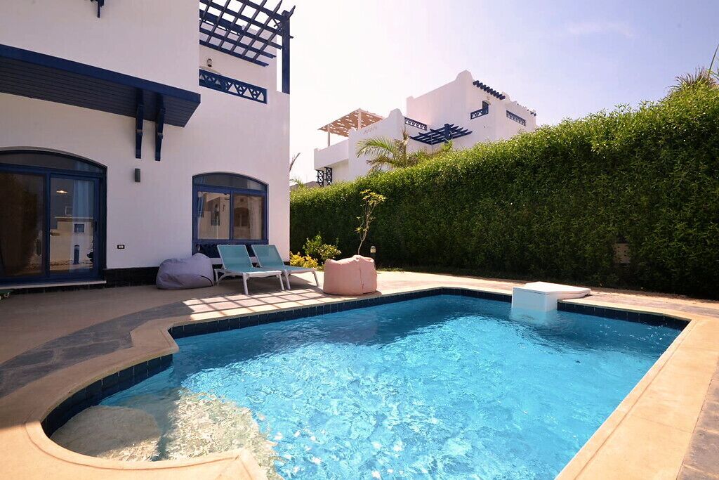 Una piscina con sillones de descanso y un edificio blanco con detalles azules.