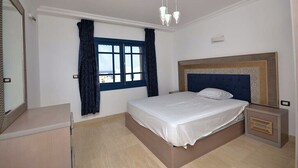 A bedroom with a bed, a bedside table, a mirror, a window with curtains, and a ceiling light fixture. - Luxury 4-Bedroom Villa for Rent in Amaros. (Red Sea Governorate)
