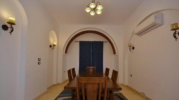 A dining area with a wooden table and chairs, an arched doorway, and wall-mounted lighting. - Luxury 4-Bedroom Villa for Rent in Amaros. (Red Sea Governorate)
