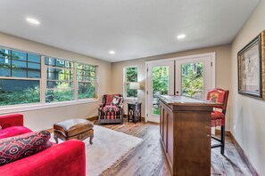 Une pièce avec une grande fenêtre, un fauteuil rouge, un bureau en bois et une petite table.