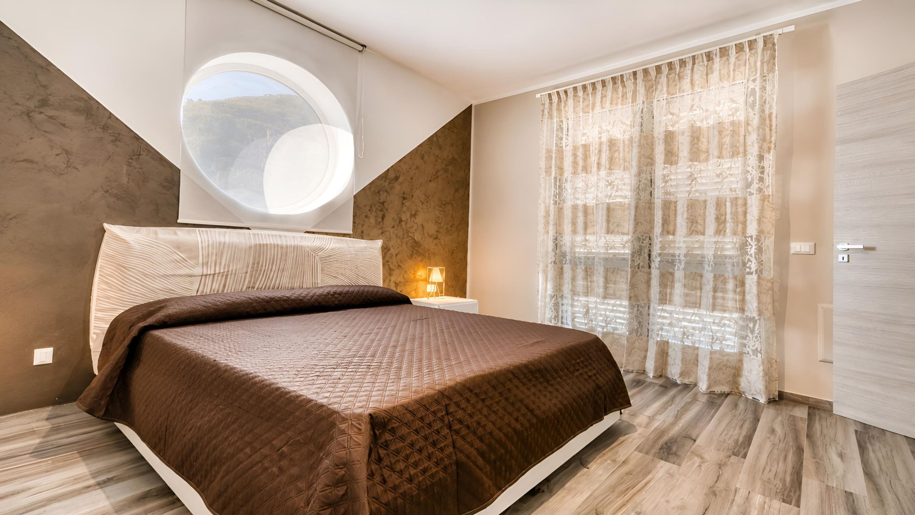 Un dormitorio con una cama grande, un tragaluz circular, una mesita de noche con lámpara y vistas al exterior a través de cortinas translúcidas.