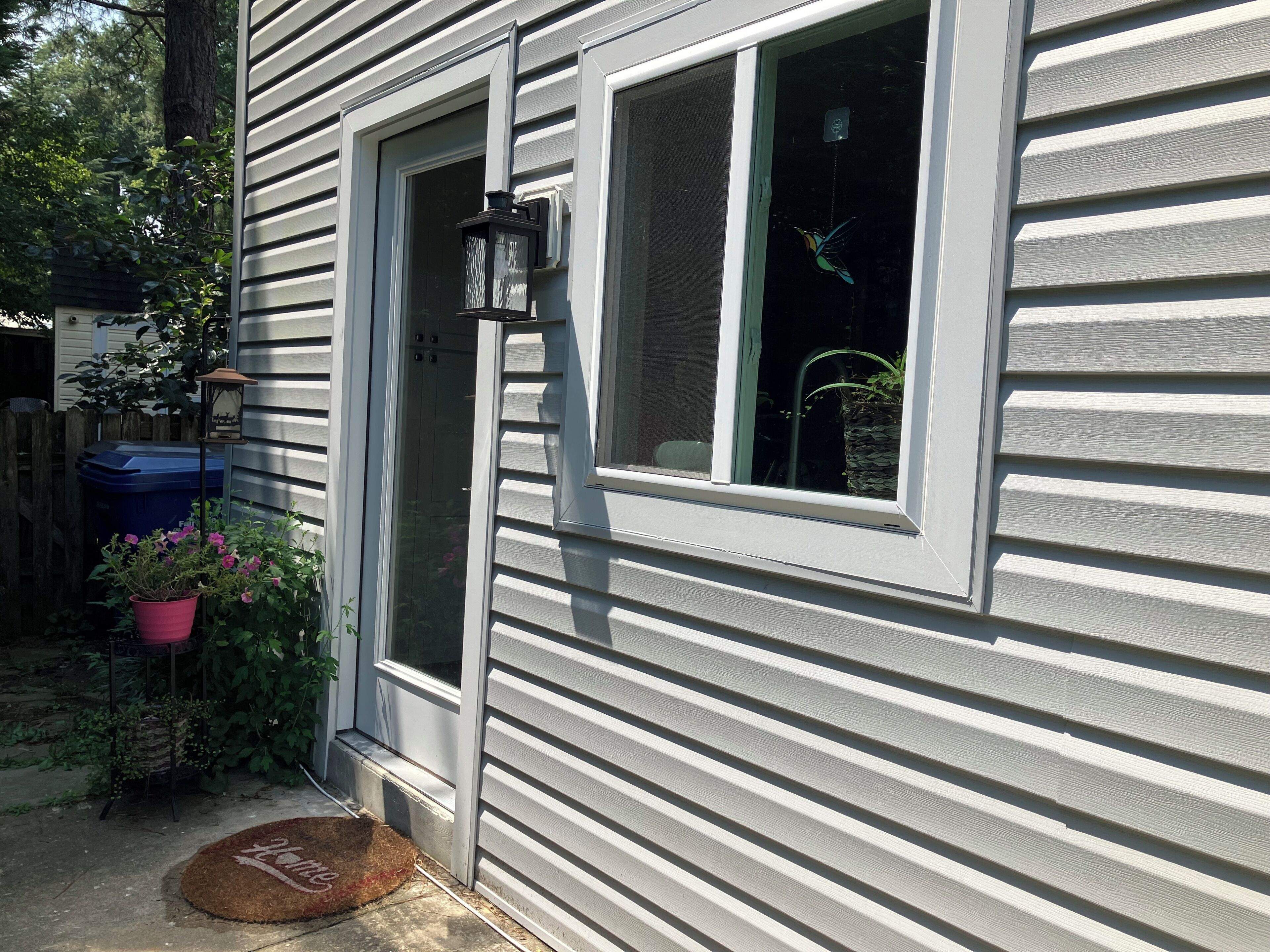 A house with a front door, a window with a bird decoration, a potted plant, and a doormat.