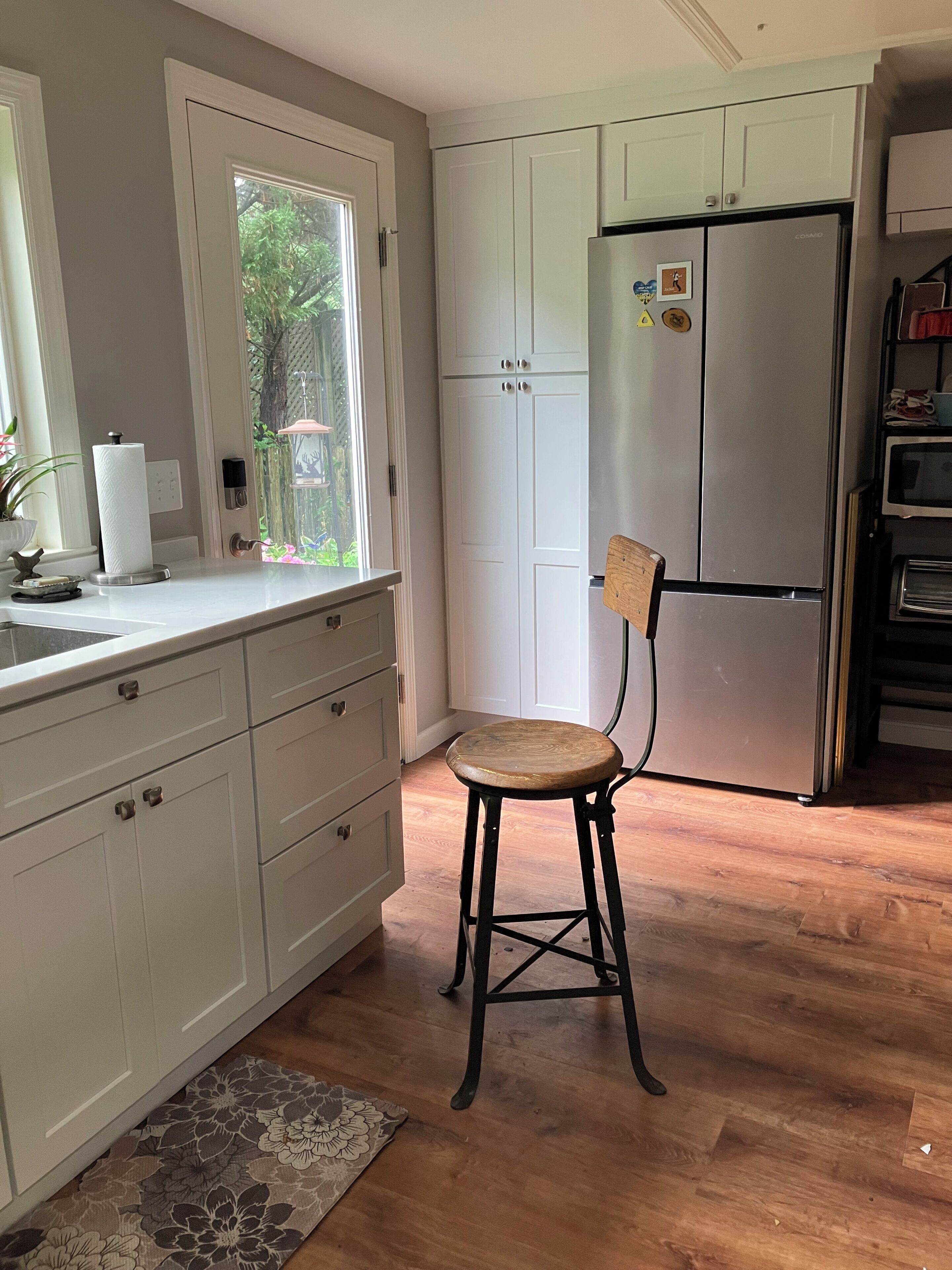 Eine Küche mit weißen Schränken, einem hölzernen Hocker, einem Kühlschrank und einem Fenster mit Blick auf Grünflächen.