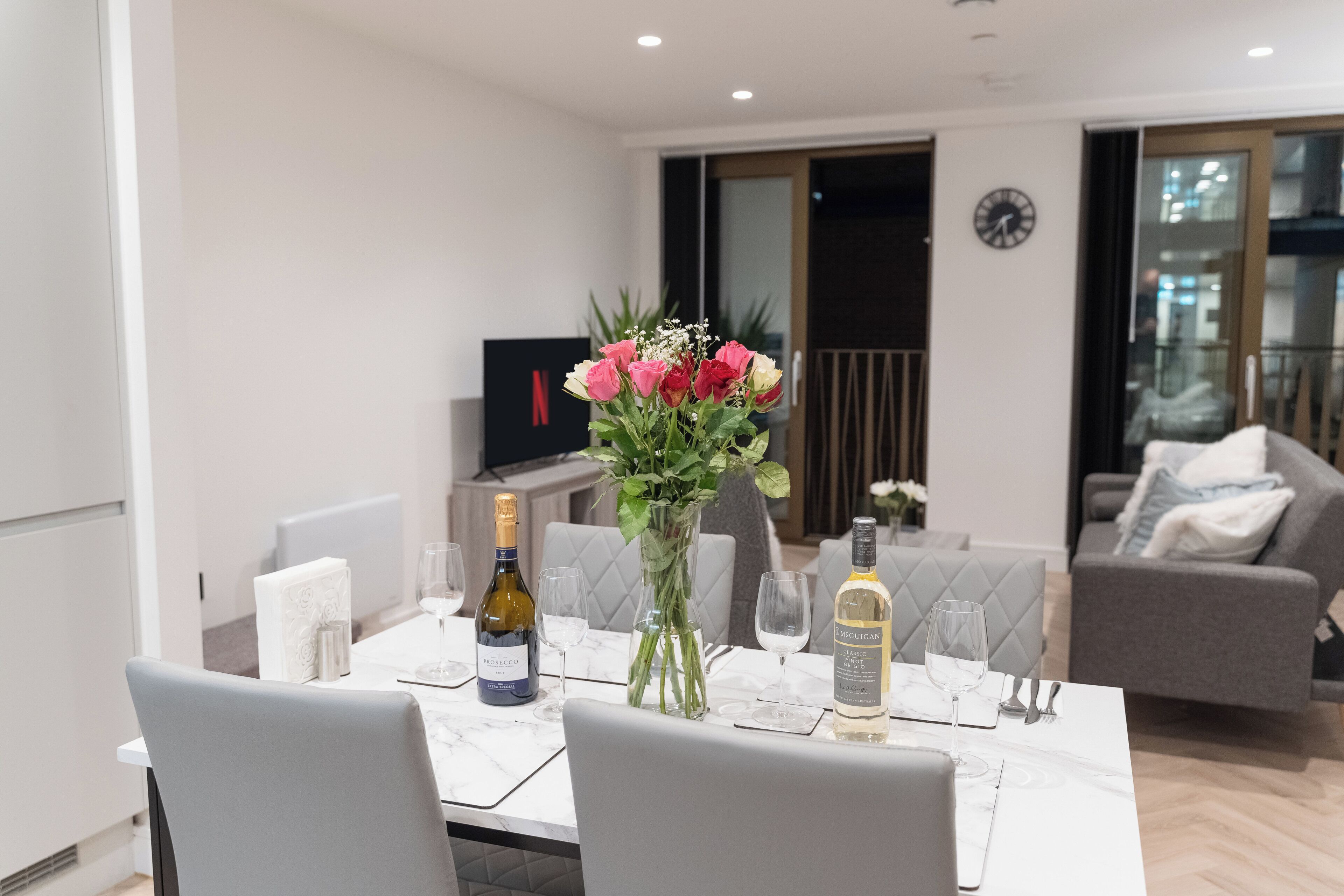 A modern dining area with a white table set for two, featuring a bottle of Prosecco and a bottle of Meiguan wine.