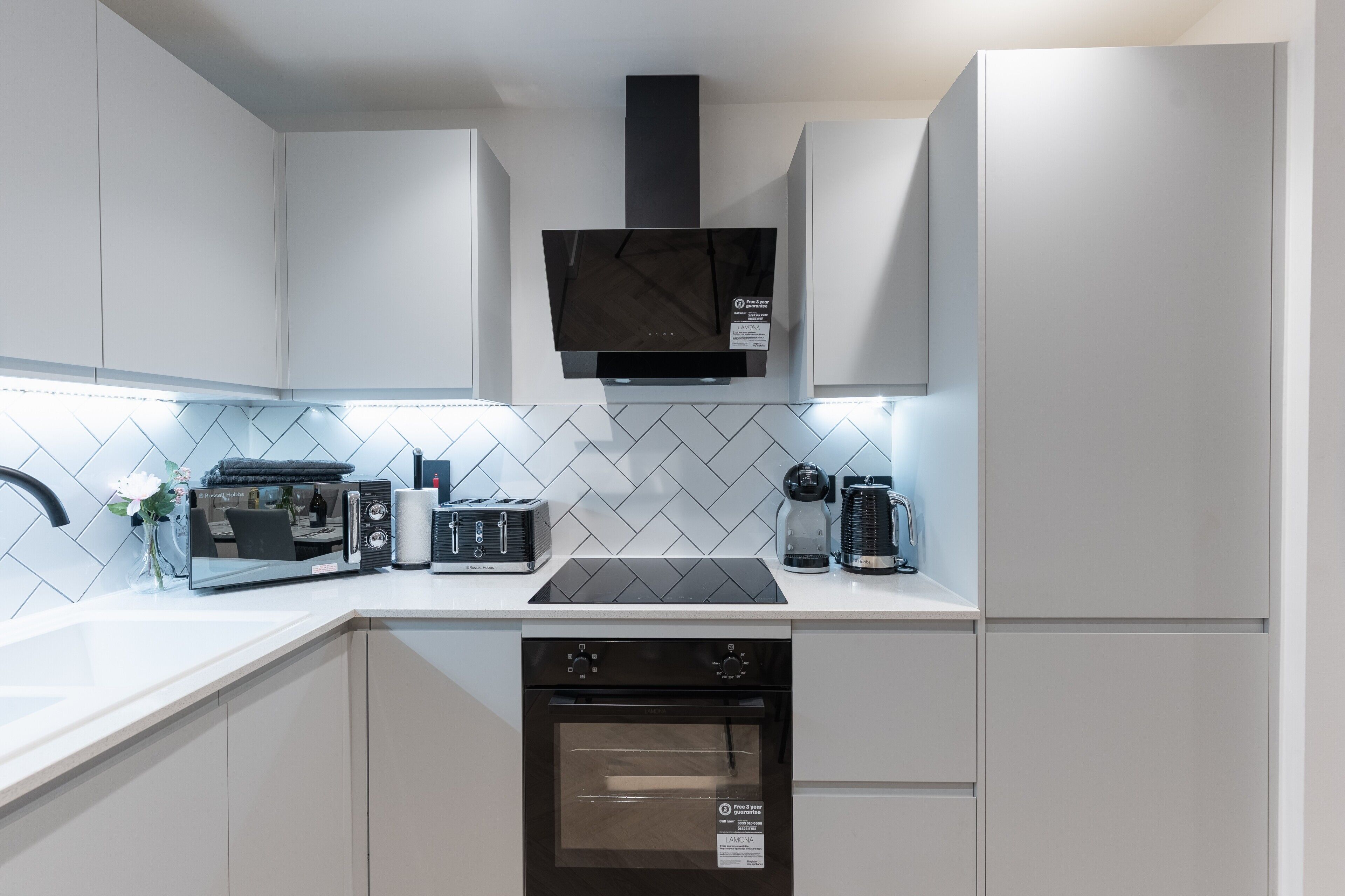 A modern kitchen with white cabinetry, a black range hood, a built-in oven, and a hob with a kettle.