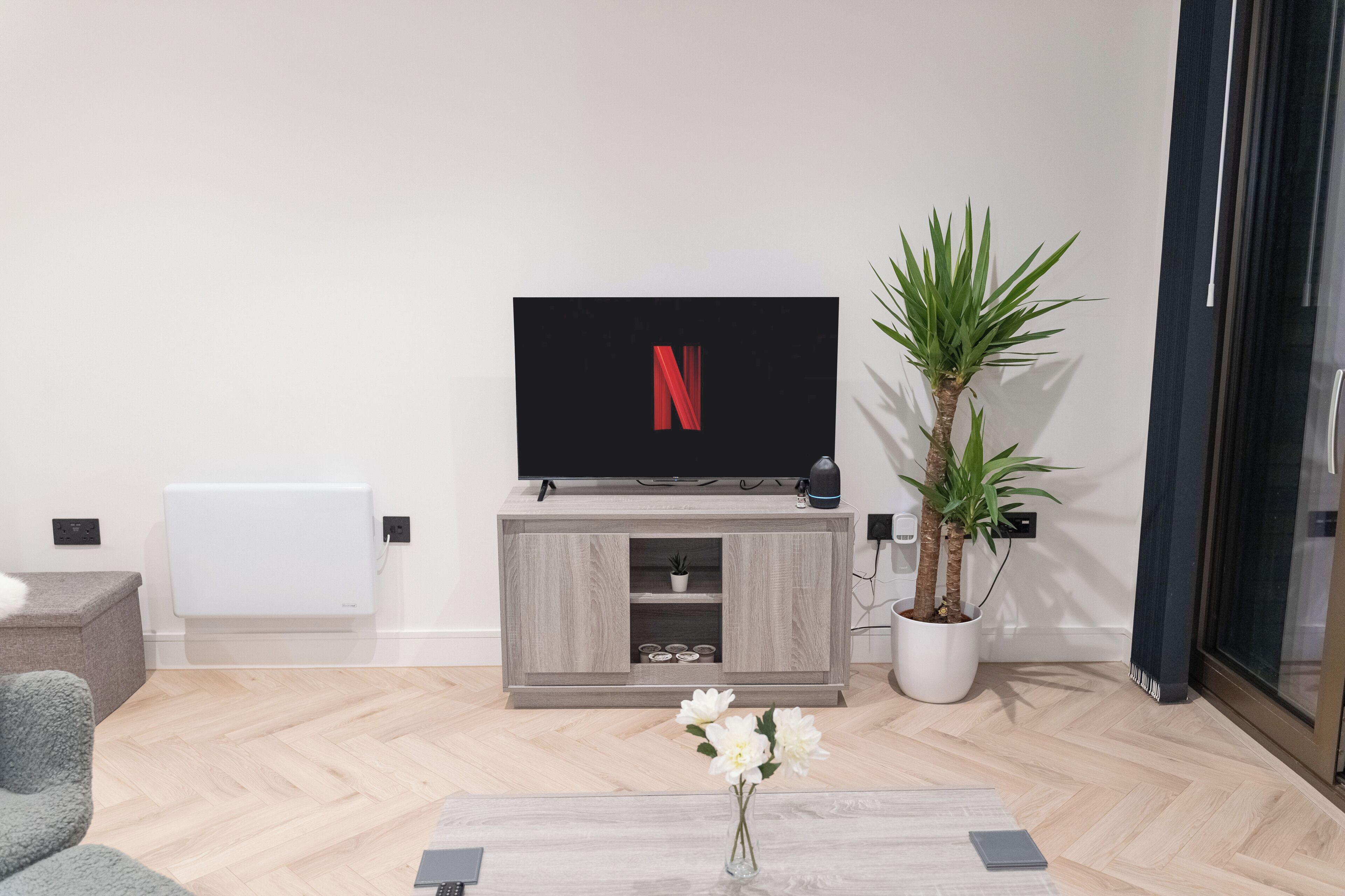 A modern living room with a flat-screen TV, a wooden entertainment unit, a potted plant, and a coffee table with a vase of flowers.
