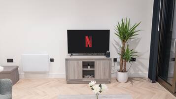 A modern living room with a flat-screen TV, a wooden entertainment unit, a potted plant, and a coffee table with a vase of flowers.
