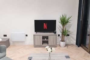 A modern living room with a flat-screen TV, a wooden entertainment unit, a potted plant, and a coffee table with a vase of flowers. - 1-Bed Flat 7 Min to Piccadilly Station (Manchester)