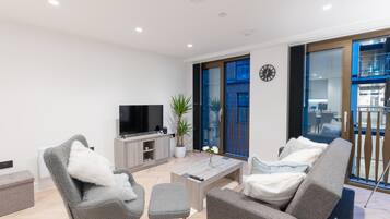 A modern living room with a flat-screen TV, grey upholstered chairs, a coffee table, and a potted plant.