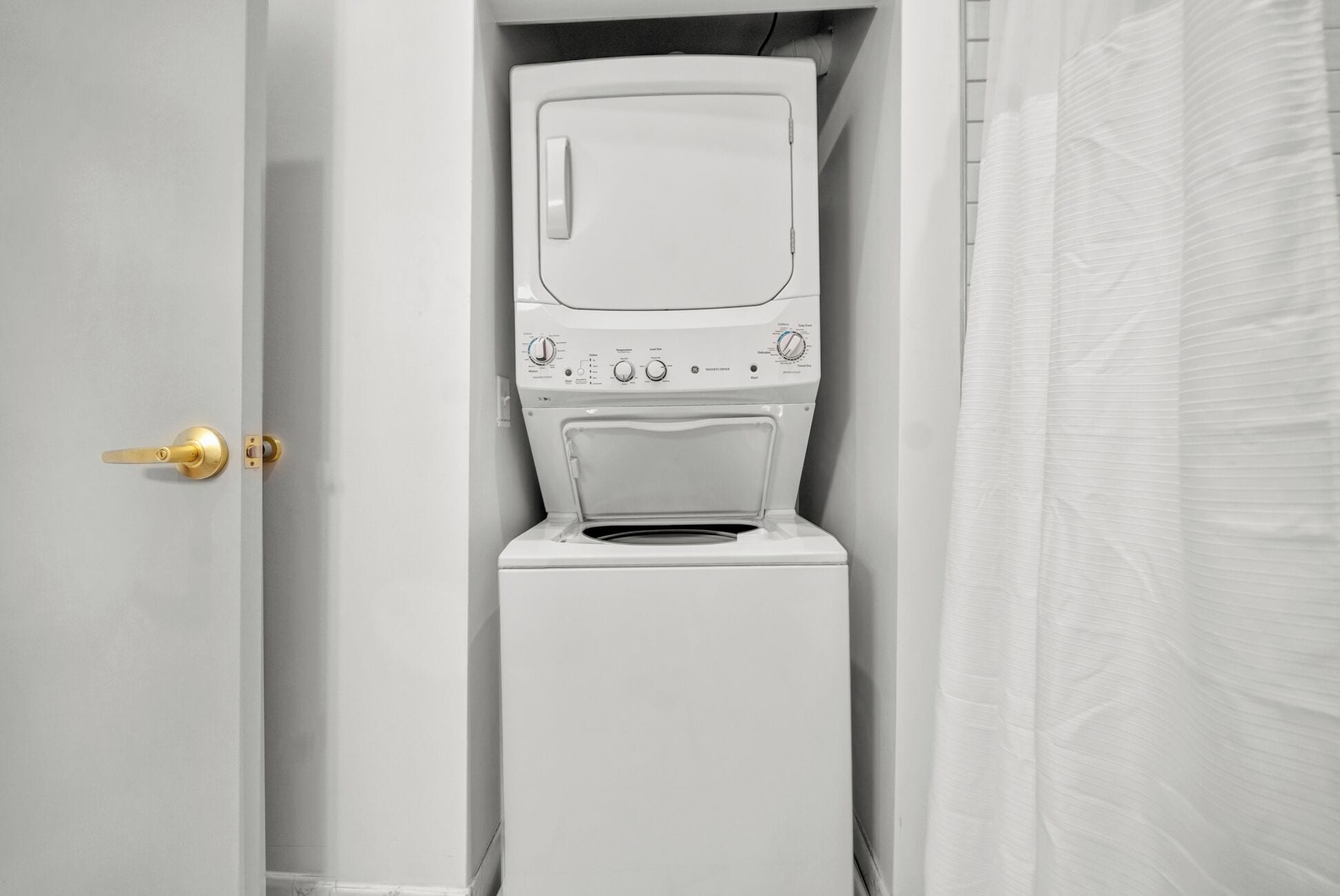 Une machine à laver blanche, dotée d’un panneau de commande, se trouve dans une salle de bain avec une porte blanche et un rideau de douche.