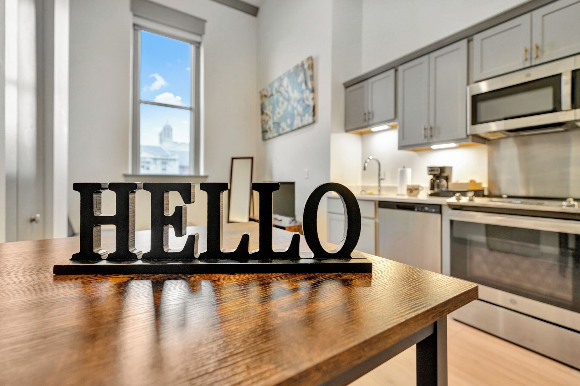 A modern kitchen with stainless steel appliances, a wooden table with a 'HELLO' sign, and a cityscape view through a window.