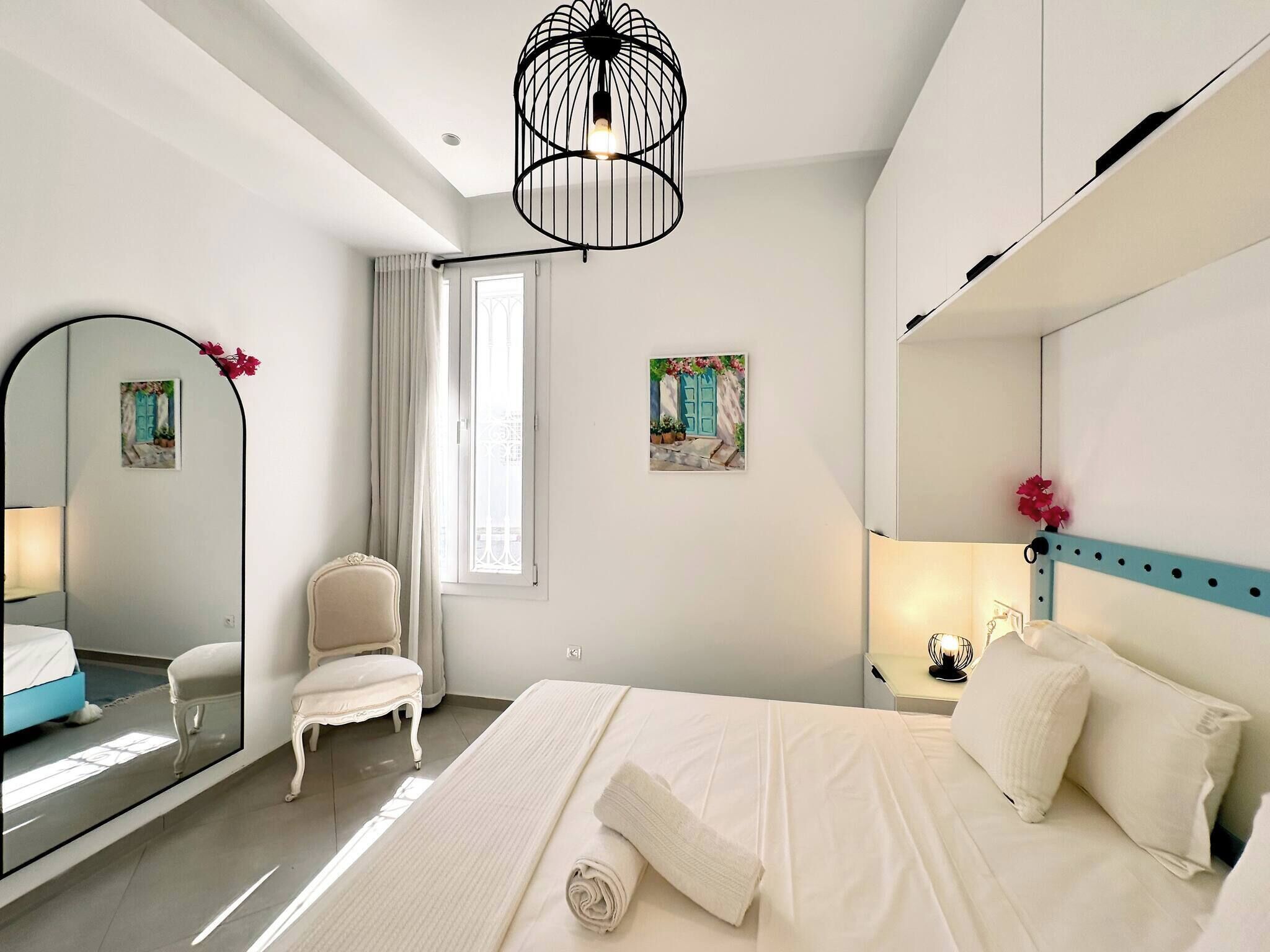 A bedroom with a bed, a chair, a mirror, a window with curtains, and a painting on the wall.