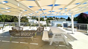 Um pátio externo com mobiliário branco, uma pérgola com painéis azulados e vista para edifícios brancos ao fundo.