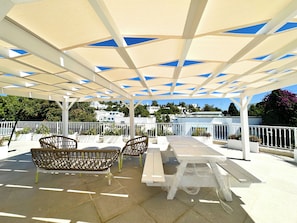 Eine Terrasse im Freien mit weißen Möbeln, einer Pergola mit blauen Paneelen und Blick auf weiße Gebäude im Hintergrund.