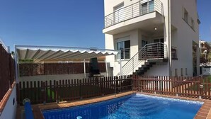 Piscina com deck de madeira, área de estar coberta e escadaria para um andar superior.
